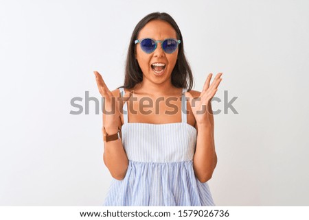 Young chinese woman wearing striped dress and sunglasses over isolated white background celebrating mad and crazy for success with arms raised and closed eyes screaming excited. Winner concept