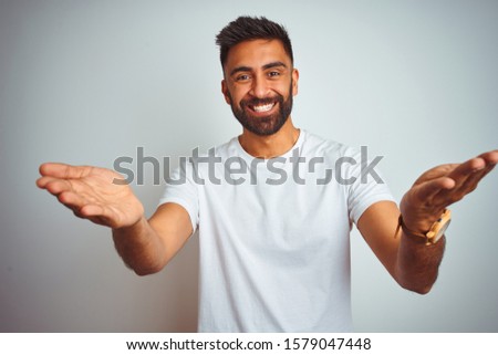 Young indian man wearing t-shirt standing over isolated white background smiling cheerful offering hands giving assistance and acceptance.