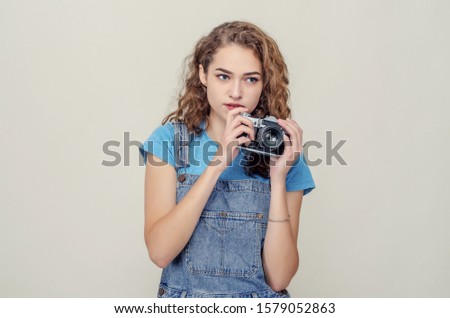 Curly cheerful brunette in denim overalls holds a film camera in her hands, having fun on the set.
