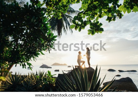The guy makes an offer to marry the girl on a large stone A man offers the hand and heart of a loved one on a tropical island. Silhouette of a couple in love in the backlight against a sunset, sea