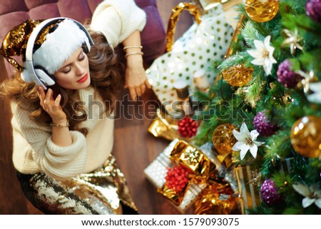 Upper view of relaxed elegant 40 year old woman with long brunette hair in gold sequin skirt and white sweater listening to the music with headphones under decorated Christmas tree near present boxes.