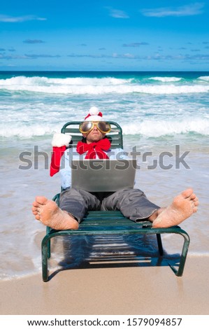 Relaxed office worker in holiday hat, sunglasses and big red bow working on his laptop on a tropical Caribbean beach