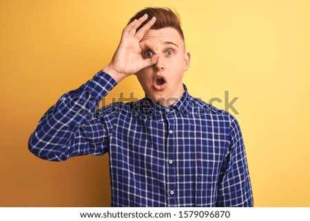 Young handsome man wearing casual shirt standing over isolated yellow background doing ok gesture shocked with surprised face, eye looking through fingers. Unbelieving expression.