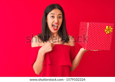 Young beautiful chinese woman holding birthday gift standing over isolated red background very happy pointing with hand and finger