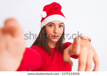 Girl wearing Christmas Santa hat make selfie by camera over isolated white background pointing with finger to the camera and to you, hand sign, positive and confident gesture from the front
