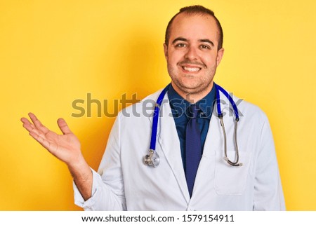 Young doctor man wearing coat and stethoscope standing over isolated yellow background smiling cheerful presenting and pointing with palm of hand looking at the camera.