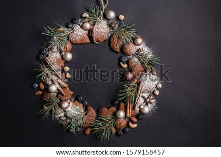 Christmas wreath of homemade cookies of various shapes, Christmas decorations, spices, nuts, blueberries and needles under powdered sugar. Black background. Top view.