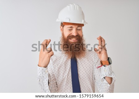 Young redhead irish architect man wearing security helmet over isolated white background gesturing finger crossed smiling with hope and eyes closed. Luck and superstitious concept.