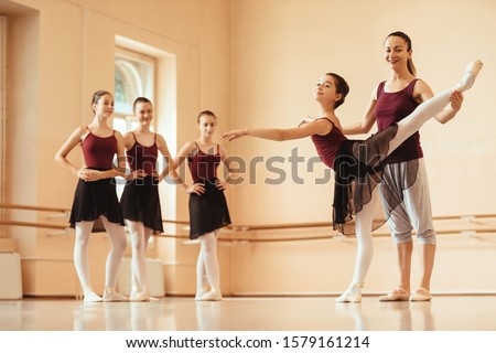 Ballet teacher helping ballerina to improve her posture while rehearsing at dance studio. Other ballerinas are standing in the background. 