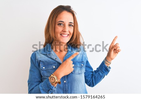 Young redhead woman wearing denim jacket standing over isolated background smiling and looking at the camera pointing with two hands and fingers to the side.