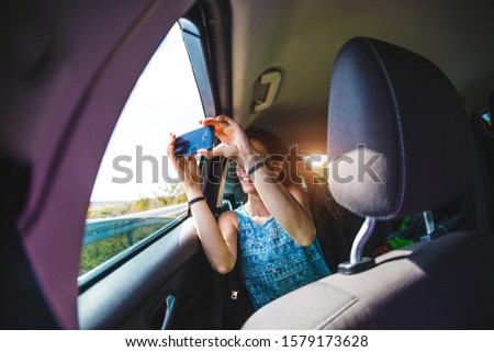 A girl sits in the back seat of a car and takes a photo of a passing landscape from a window. A woman is being photographed in the back seat of a vehicle. Smiling brunette takes a selfie.