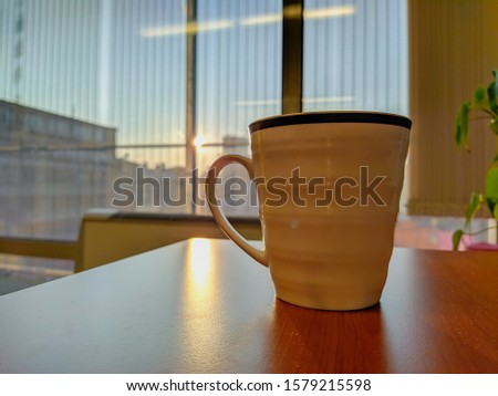 Closeup of white mug cup made of porcelain ceramics placed on a wooden desk with beautiful view of outside through the mirror while at a job