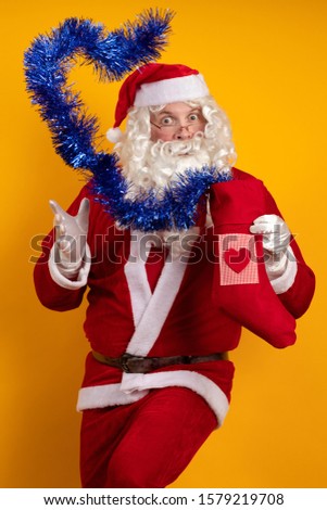 Santa Claus with a long beard and glasses holds in his hands a Christmas sock with gifts and a blue garland of tinsel and poses on a yellow background
