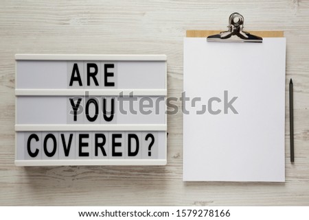 Top view, 'Are you covered?' words on a modern board, clipboard with blank sheet of paper on a white wooden surface. Overhead, from above, flat lay. 