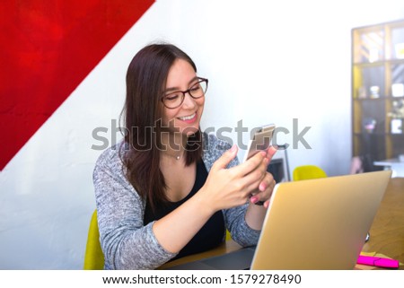 Happy smiling female in glasses skilled content writer reading e-mail on mobile phone while sitting at workplaces in co-working space. Office worker online chatting on cellphone during webinar 