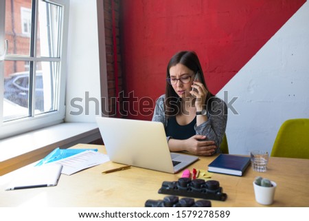 Angry upset female in glasses skilled business woman listening about bad news via mobile phone conversation and watching on laptop computer screen while sitting at workplace in office interior 