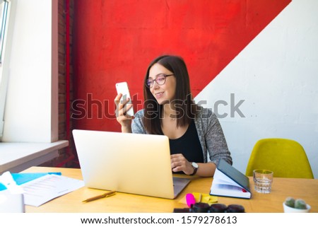 Happy smiling hipster girl in glasses successful marketing specialist using messenger on mobile phone while sitting at table with laptop computer in modern co-working space 