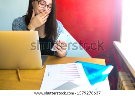 Female in spectacles professional financier reading good text message on cellphone while sitting at desktop with laptop computer near copy space for promotional content 