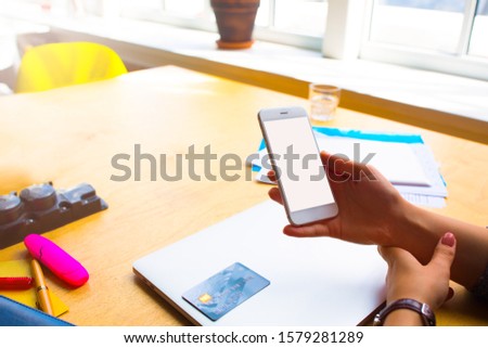 Woman financier online booking via cellphone with blank screen and using for payment credit debit card while sitting at desktop with closed netbook in modern office interior 