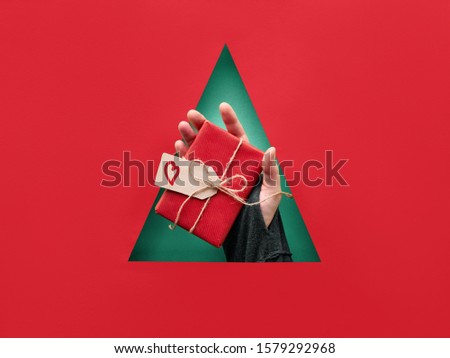 Flame scarpet paper Xmas background with triangular hole in the middle of red paper. Hand holding wrapped Christmas gift with heart on cardboard tag on green background. Happy Holidays!