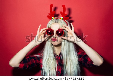 Portrait of happy young blonde girl with Christmas balls on eyes, wearing deer horns, isolated on red background.