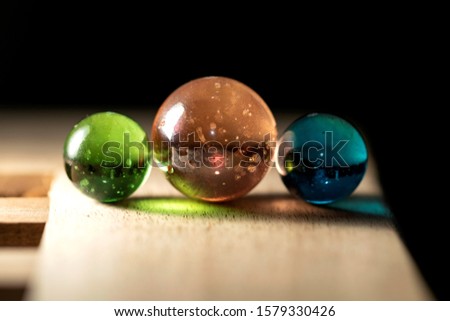 A close up portrait of three different colored marbles on a wooden surface casting colorful reflections. The glass spheres are orange, green and blue.