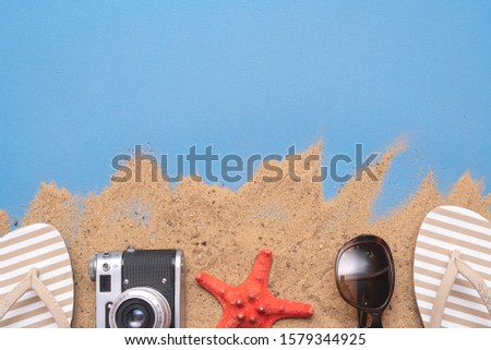 Flip flops shoes, sunglasses , retro photo camera and red starfish on the sand on blue flat lay background with copy space.