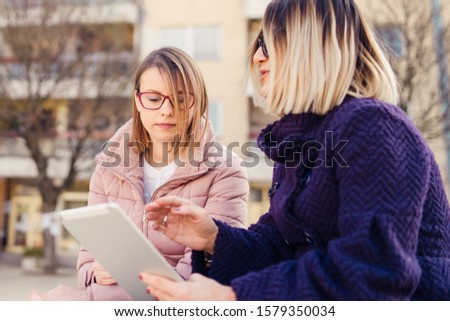 Two women female friends using digital tablet notebook in the city looking to the pictures or map application project in winter or autumn day talking discussing work