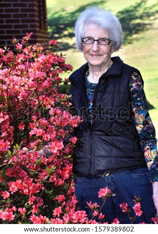 Grandmother stands and admires her pink azalea bush.  It is in full bloom.  She is wearing a black vest and jeans.