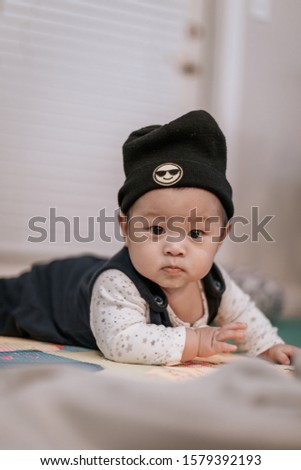 Asian baby boy wearing black knitted beanie lying on play mat during tummy time at home. Child is 6 months old.