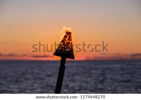 A silhouette of a gas torch with flame around it against scenic sunset. Red sun going down into the ocean, line of clouds above the horizon., Big Island, Hawaii