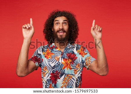 Studio shot of positive young brunette curly man with beard showing up with index fingers while posing against red background in multi-colored flowered shirt
