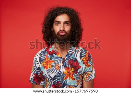 Funny portrait of young dark haired curly man with beard making ridiculous faces while posing against red background, wearing multi-colored flowered shirt