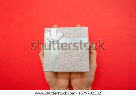 Top view of women's hands holding a shiny gray gift box with a white bow on a red background, copy space