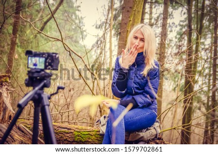 blonde girl in a blue jacket and jeans, boots writes a blog, as if a blogger is sitting in the forest, sitting on a fallen tree, smiling, blowing a kiss to the camera.. Writes a blog on DSLR.