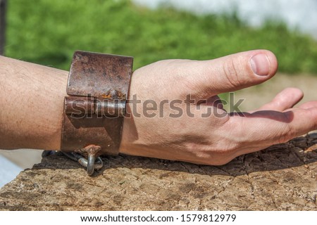 The man’s hand is shackled in metal medieval ancient handcuffs on a tree cut, on a stump. Punishment. Chained Man