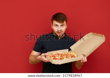 A funny man with a beard holds a pizza box in delivery and looks into the camera with a hungry lustful face. Portrait of hungry man with pizza box on red background. Fast junk food concept. Royalty-Free Stock Photo #1579818967