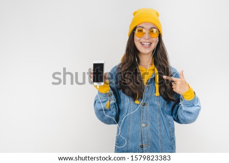 Portrait excited girl in hat and earphones pointing finger at blank screen mobile phone over white background