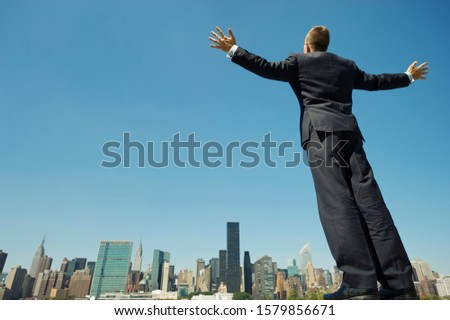 Full length view of powerful businessman standing tall with his arms spread above the city skyline under bright blue sky copy space