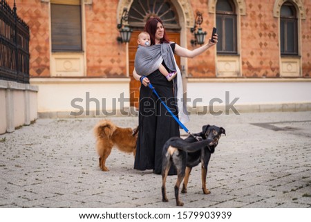 A young woman with a baby in a sling walking her dogs in city and taking photo with mobile phone
