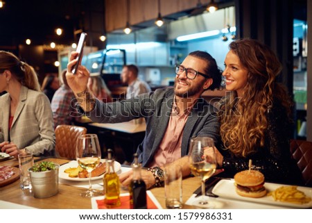 Cute caucasian smiling couple in love sitting in restaurant and taking selfie. Around them are their friends chatting and having dinner.