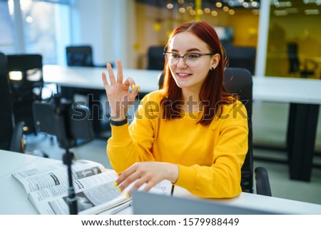 Female blogger with laptop recording video while sitting at office and talking  about startup small business. Teenager student in a yellow sweater having fun vlogging live feeds on social media.