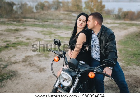 Stylish man biker and gorgeous brunette girl in a black dress and cuddling tenderly on an expensive black motorcycle. Portrait of a couple in love on the nature. Photography, concept, love story.