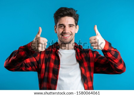 Positive young man smiles to camera. Hipster guy showing thumb up sign over blue background. Winner. Success. Body language.
