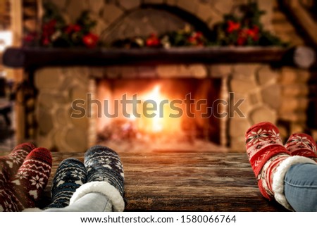 Wooden retro table and free space for your decoration.People legs with christmas socks.Blurred fireplace with xmas decoration.Warm orange fire and dark mood interior of home. 