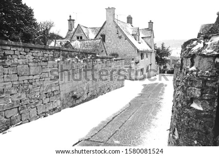 A snow covered black and white image of a winter scene looking down a narrow road in the village of Painswick, The Cotswolds, Gloucestershire, UK