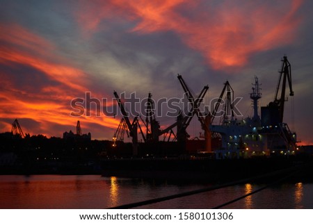 Colorful sunset over sea port and industrial cranes. Bright sunset in seaport. Large silhouettes of cargo cranes. Beautiful landscape with fiery sunset sky and sea.