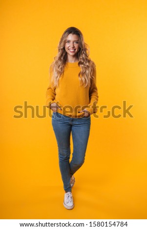 Full length image of lovely blonde woman with long hair wearing warm sweater smiling at camera isolated over yellow background