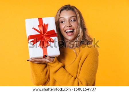 Image of satisfied blonde woman in warm sweater smiling and holding present box isolated over yellow background