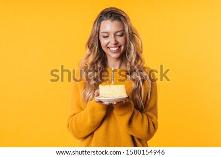 Image of cheerful blonde woman in warm sweater smiling and holding birthday cake with candle isolated over yellow background